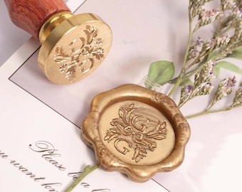 May include: A gold-colored wax seal stamp with a wooden handle and a floral design with the letter "G". A matching wax seal is also present. The stamp and seal are on a white surface with text.