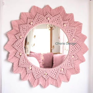 May include: A circular wall mirror framed by dusty pink macrame cord arranged in a sunburst pattern with pointed edges. The design features intricate knotted textures and small, light-coloured wooden beads integrated into the frame. The text Cherie Design is visible on the mirror surface.