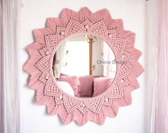 May include: A circular wall mirror framed by dusty pink macrame cord arranged in a sunburst pattern with pointed edges. The design features intricate knotted textures and small, light-colored wooden beads integrated into the frame. The text Cherie Design is visible on the mirror surface.