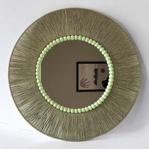 Op de afbeelding: Een ronde wandspiegel met een brede lijst omwikkeld met getextureerd beige koord. De binnenrand van de lijst is versierd met een ring van kleine, licht limoengroene bolvormige kralen. Het spiegeloppervlak toont de reflectie van een abstracte kunstprint.