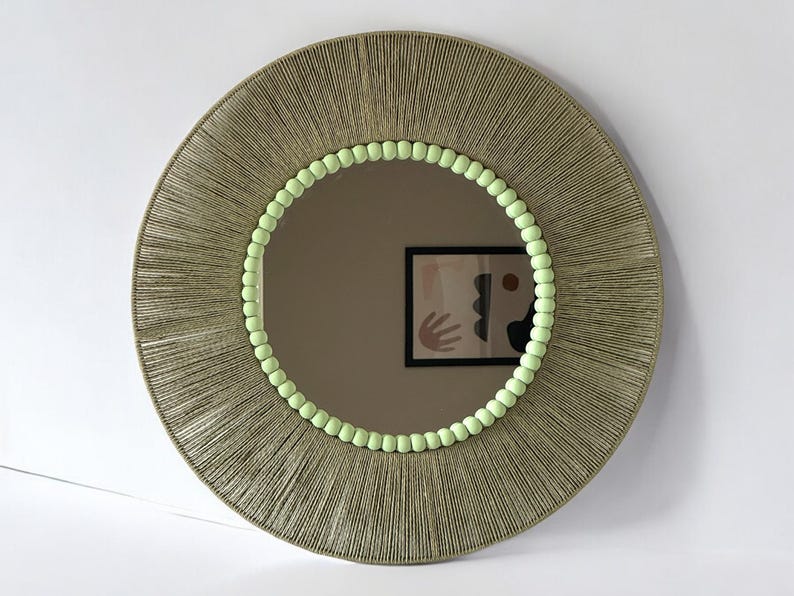 Op de afbeelding: Een ronde wandspiegel met een brede lijst omwikkeld met getextureerd beige koord. De binnenrand van de lijst is versierd met een ring van kleine, licht limoengroene bolvormige kralen. Het spiegeloppervlak toont de reflectie van een abstracte kunstprint.