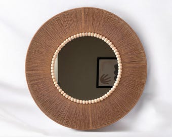 Könnte beinhalten: Ein runder Wandspiegel mit einem Rahmen, der mit beigefarbener Jutekordel umwickelt ist. Der innere Rand des Rahmens ist mit einer Reihe kleiner, heller Holzperlen verziert, die die reflektierende Glasfläche des runden Spiegels umgeben.