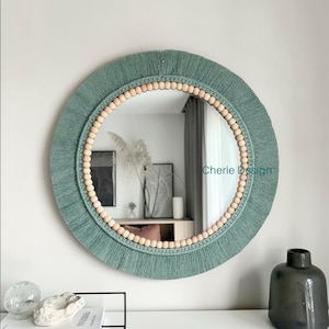 Peut inclure: Un miroir mural rond doté d'une bordure à franges en fibre vert sauge et d'un cadre circulaire composé de perles en bois clair. Le texte Cherie Design est visible sur la droite de la surface du miroir, reflétant l'intérieur d'une pièce.