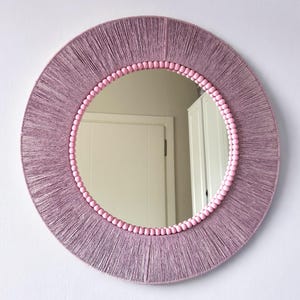 Op de afbeelding: Een ronde wandspiegel met een brede lijst omwikkeld met mauvekleurig garen. De binnenrand van de lijst is versierd met een rij kleine, lichtroze bolvormige kralen die het reflecterende glazen oppervlak omlijsten.