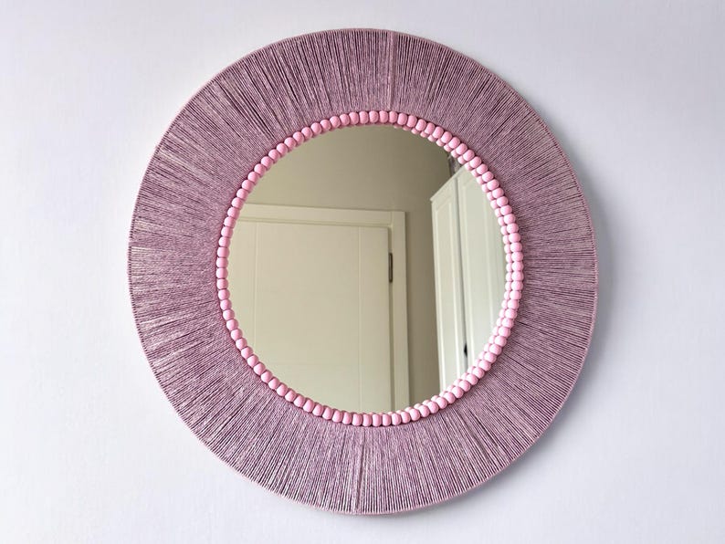 Op de afbeelding: Een ronde wandspiegel met een brede lijst omwikkeld met mauvekleurig garen. De binnenrand van de lijst is versierd met een rij kleine, lichtroze bolvormige kralen die het reflecterende glazen oppervlak omlijsten.