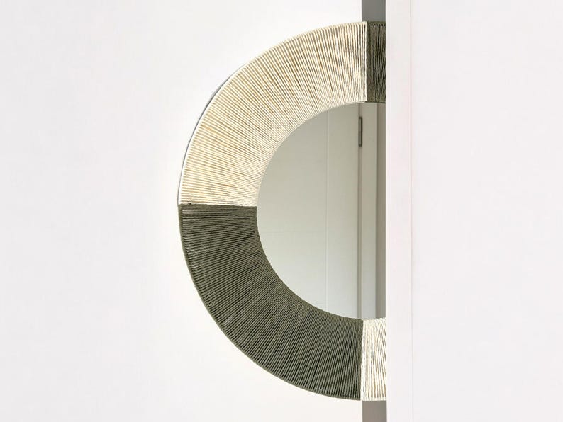Pode incluir: Um espelho de parede semicircular com uma moldura envolta em segmentos alternados de fio creme e cinza escuro. O espelho tem uma borda vertical plana e uma borda externa curva, com o fio criando um padr&atilde;o radial texturizado ao redor.