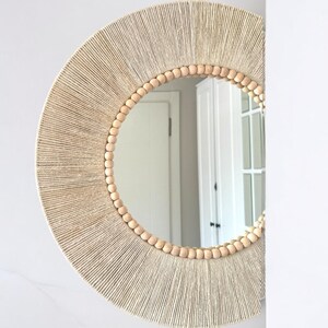 May include: A circular wall mirror featuring a frame wrapped in light beige natural fiber twine. The inner edge of the frame is decorated with a single row of smooth, round, light-colored wooden beads that encircle the reflective glass surface.