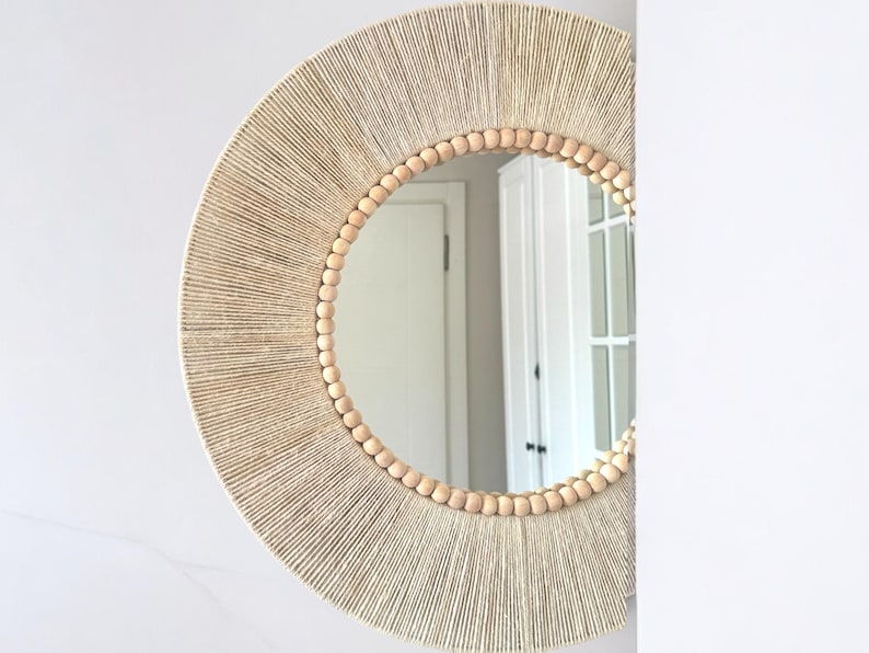 May include: A circular wall mirror featuring a frame wrapped in light beige natural fiber twine. The inner edge of the frame is decorated with a single row of smooth, round, light-colored wooden beads that encircle the reflective glass surface.