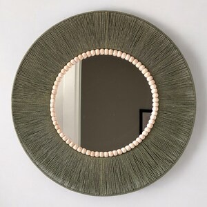 May include: A circular wall mirror featuring a frame wrapped in olive green cord. The inner edge of the frame is decorated with a single row of small, light-colored wooden beads that surround the reflective glass surface of the mirror.