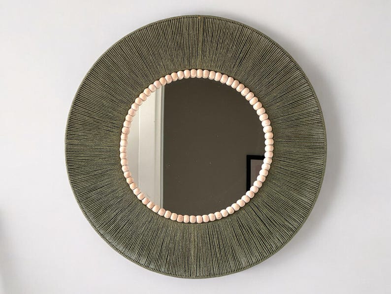 May include: A circular wall mirror featuring a frame wrapped in olive green cord. The inner edge of the frame is decorated with a single row of small, light-colored wooden beads that surround the reflective glass surface of the mirror.