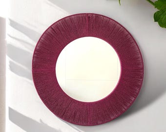 May include: A circular wall mirror featuring a wide, deep magenta frame wrapped in tightly wound, thin cord or thread. The central glass surface is clear and reflective, showing a subtle horizontal line reflection within the circular frame.