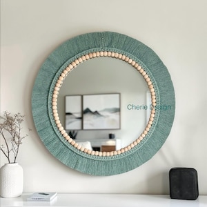 May include: A round wall mirror featuring a sage green fibre fringe border and an inner ring of light-coloured wooden beads. The text Cherie Design is visible on the mirror surface. The mirror is mounted on a wall above a white cabinet.