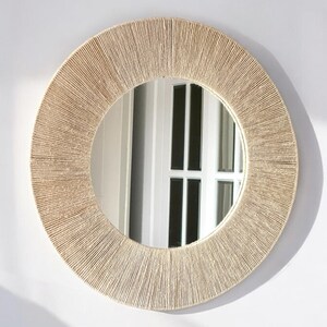 May include: A circular wall mirror featuring a wide frame wrapped in tightly coiled, light beige natural fiber cord. The reflective glass center provides a clear view, while the textured, radial pattern of the cord creates a distinct border.