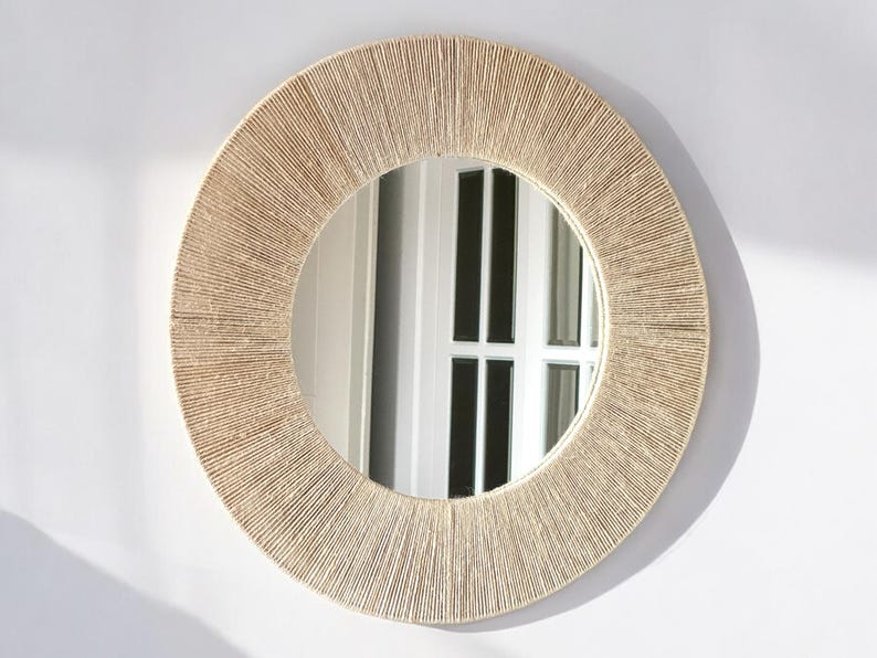 May include: A circular wall mirror featuring a wide frame wrapped in tightly coiled, light beige natural fiber cord. The reflective glass center provides a clear view, while the textured, radial pattern of the cord creates a distinct border.