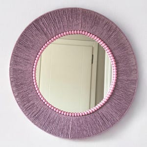 Op de afbeelding: Een ronde wandspiegel met een frame omwikkeld met mauvekleurig garen. De binnenrand van het frame is versierd met een ring van kleine, lichtroze bolvormige kralen die het reflecterende glasoppervlak van de ronde spiegel omringen.