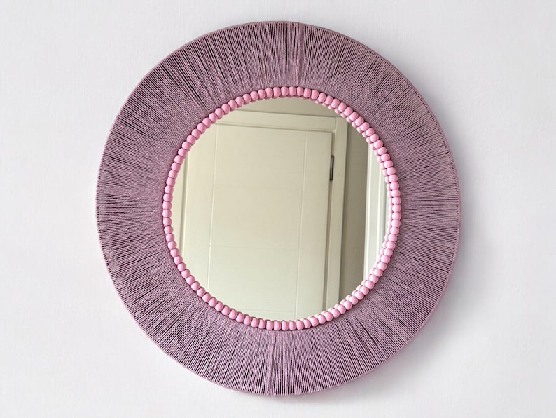 Op de afbeelding: Een ronde wandspiegel met een frame omwikkeld met mauvekleurig garen. De binnenrand van het frame is versierd met een ring van kleine, lichtroze bolvormige kralen die het reflecterende glasoppervlak van de ronde spiegel omringen.