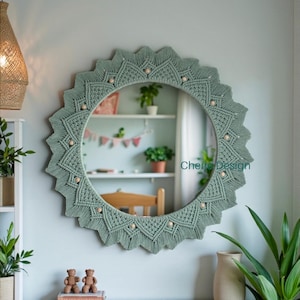 Può includere: Uno specchio da parete rotondo con cornice in macramè verde salvia dal design a raggiera, impreziosito da piccole perline di legno. Lo specchio riporta la scritta Cherie Design in lettere color ottanio. L'intricato motivo a nodi crea un bordo stellato.