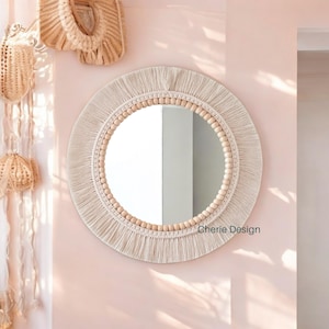 Puede incluir: Un espejo de pared redondo con un marco decorado con flecos de macramé de color crema y un borde de cuentas de madera clara. El texto Cherie Design está impreso en verde oscuro en la parte inferior derecha del marco del espejo.