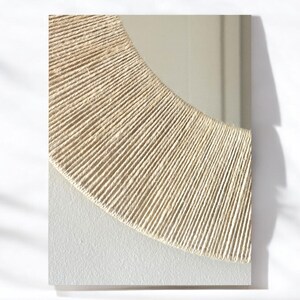 May include: A rectangular mirror featuring a semi-circular frame section wrapped in natural beige jute twine. The lower portion of the mirror surface is framed by a textured, off-white matte border that contrasts with the reflective glass.