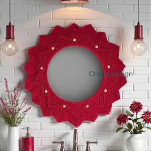 May include: A round red macrame mirror with a scalloped edge and wooden bead accents. The mirror hangs above a white sink with a chrome faucet. The background is a white brick wall, with red pendant lights and decorative vases.