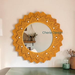 May include: A round wall mirror featuring a mustard yellow macrame border with a sunburst pattern, accented by small white wooden beads. The text Cherie Design is visible on the mirror surface, which reflects a room interior with a wooden shelf.