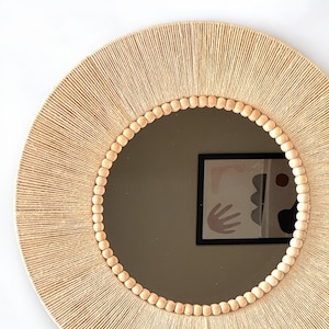 May include: A round wall mirror featuring a frame wrapped in light beige natural fiber cord. The inner edge of the frame is decorated with a single row of small, smooth, light-colored wooden beads that encircle the reflective glass surface.