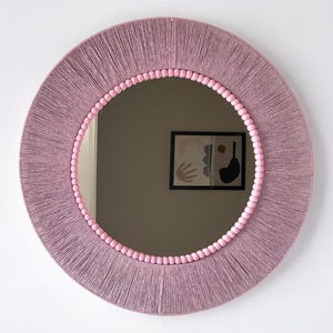 Op de afbeelding: Een ronde spiegel met een frame omwikkeld met mauvekleurig garen. De binnenrand van het frame is versierd met een ring van kleine, lichtroze bolvormige kralen. Het spiegeloppervlak toont de reflectie van een ingelijst abstract kunstwerk aan de muur.
