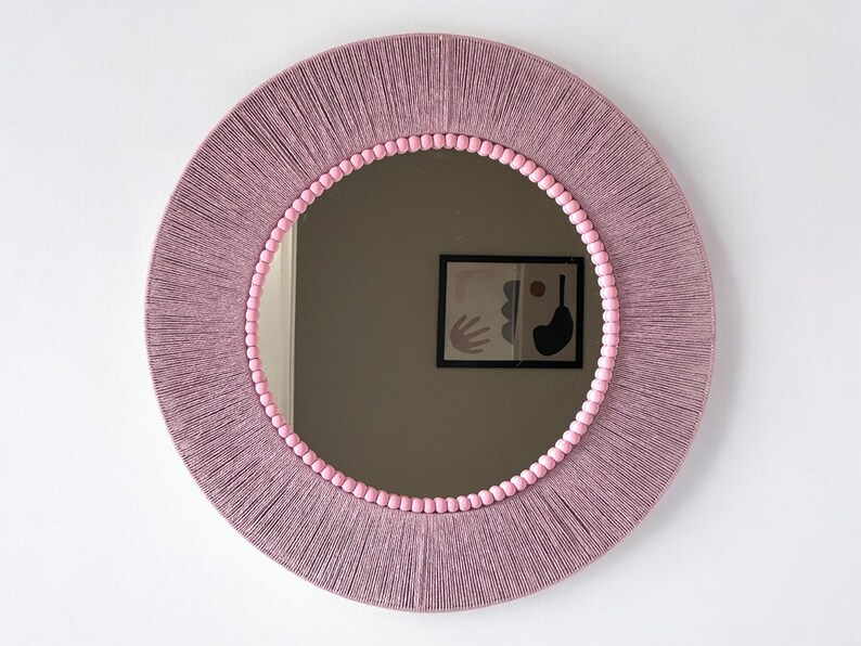Op de afbeelding: Een ronde spiegel met een frame omwikkeld met mauvekleurig garen. De binnenrand van het frame is versierd met een ring van kleine, lichtroze bolvormige kralen. Het spiegeloppervlak toont de reflectie van een ingelijst abstract kunstwerk aan de muur.