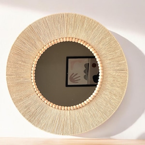May include: A round wall mirror featuring a frame wrapped in light beige natural fiber cord. The inner edge of the frame is decorated with a single row of smooth, round, light-colored wooden beads that encircle the circular glass mirror surface.