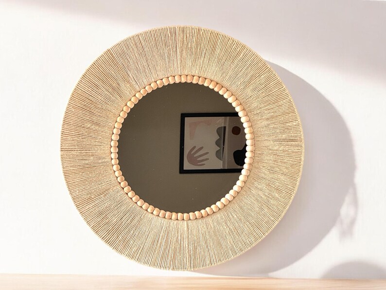 May include: A round wall mirror featuring a frame wrapped in light beige natural fiber cord. The inner edge of the frame is decorated with a single row of smooth, round, light-colored wooden beads that encircle the circular glass mirror surface.
