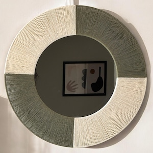 Pode incluir: Um espelho de parede circular com uma moldura envolta em segmentos alternados de fio na cor creme e verde s&aacute;lvia. O fio &eacute; enrolado firmemente em um padr&atilde;o radial ao redor de toda a circunfer&ecirc;ncia do espelho, criando um aspecto linear.