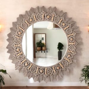 May include: A round wall mirror featuring a decorative frame made of light gray macrame cord and natural wooden beads. The frame has a sunburst design with intricate knotted patterns and fringe edges surrounding the circular glass center.