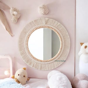 May include: A round wall mirror featuring a frame decorated with natural wooden beads and cream-colored macrame fringe. The mirror surface reflects a room interior, and the text Cherie Design is printed in small black letters on the glass.