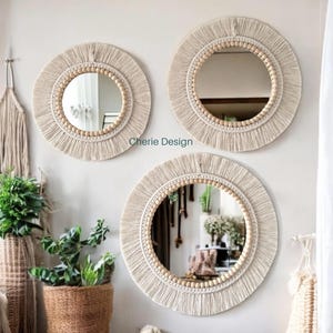 Pode incluir: Três espelhos de parede circulares com bordas de franjas de macramé na cor creme e anéis internos de contas de madeira natural. O texto Cherie Design é visível entre os espelhos. Cada espelho possui um painel de vidro central que reflete.