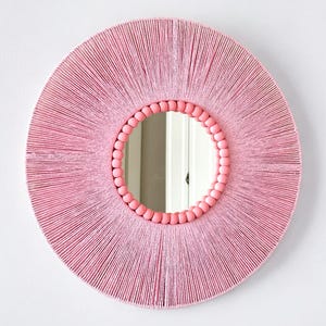 Op de afbeelding: Een ronde wandspiegel met een frame dat is omwikkeld met lichtroze draad, wat een stralend patroon cre&euml;ert. De binnenrand van het frame is versierd met een enkele rij gladde, ronde, lichtroze houten kralen die het spiegelglas omringen.