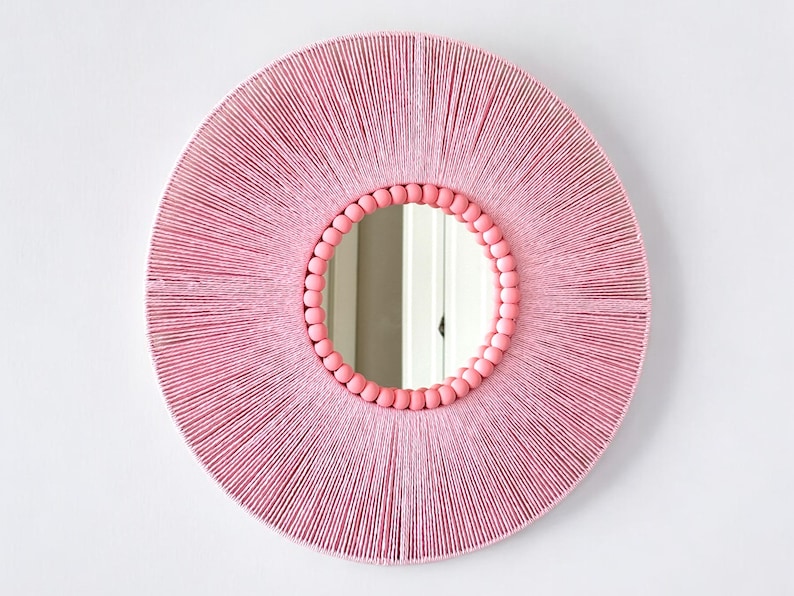 Op de afbeelding: Een ronde wandspiegel met een frame dat is omwikkeld met lichtroze draad, wat een stralend patroon cre&euml;ert. De binnenrand van het frame is versierd met een enkele rij gladde, ronde, lichtroze houten kralen die het spiegelglas omringen.