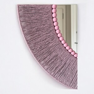Op de afbeelding: Een halfronde wandspiegel met een frame omwikkeld met getextureerd mauve garen. De binnenrand van de spiegel is bekleed met een rij gladde, ronde, lichtroze houten kralen die de welving van het glazen oppervlak volgen.
