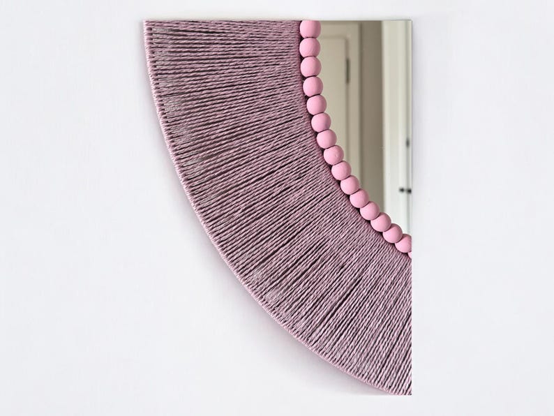 Op de afbeelding: Een halfronde wandspiegel met een frame omwikkeld met getextureerd mauve garen. De binnenrand van de spiegel is bekleed met een rij gladde, ronde, lichtroze houten kralen die de welving van het glazen oppervlak volgen.