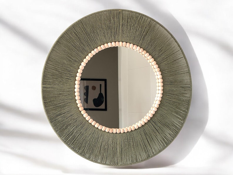 May include: A round wall mirror featuring a wide frame wrapped in olive green thread. The inner edge of the frame is decorated with a single row of small, light-colored wooden beads that encircle the reflective glass surface of the mirror.