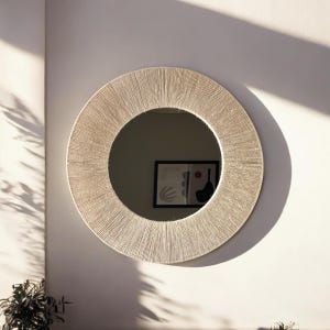 May include: A circular wall mirror featuring a wide frame wrapped in light beige natural fiber cord. The mirror surface reflects a framed abstract art piece with dark shapes on a light background, creating a clean and textured appearance.
