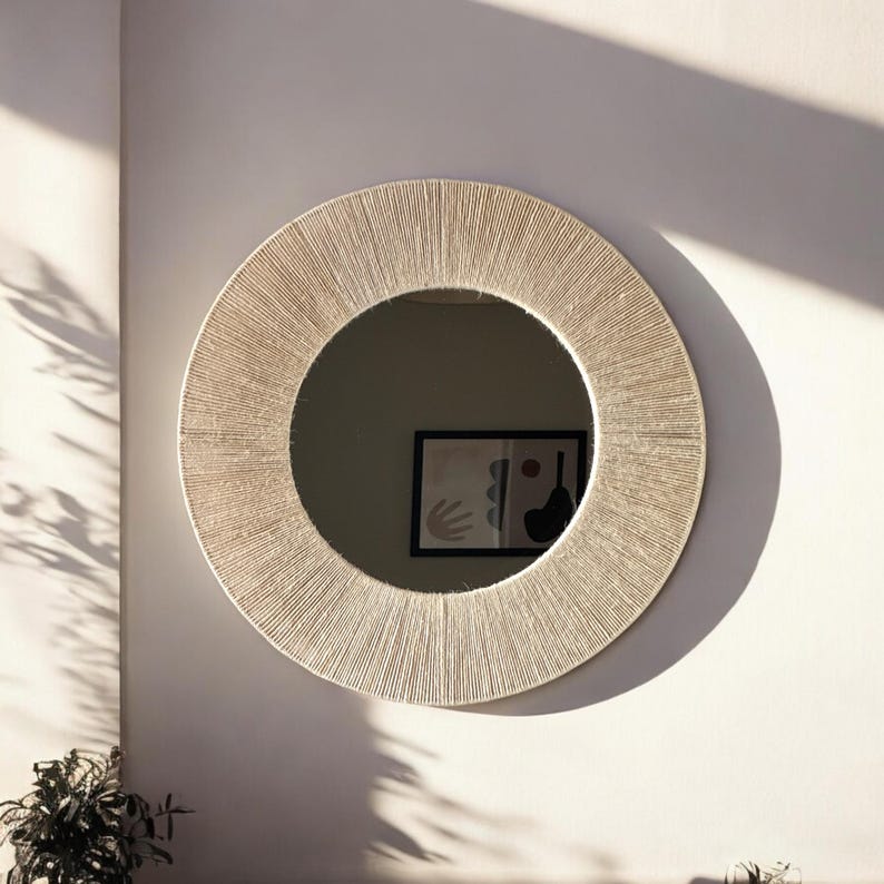 May include: A circular wall mirror featuring a wide frame wrapped in light beige natural fiber cord. The mirror surface reflects a framed abstract art piece with dark shapes on a light background, creating a clean and textured appearance.