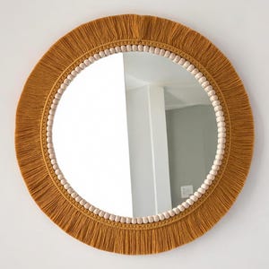 May include: A circular wall mirror featuring a decorative border of light-colored wooden beads and a wide, mustard-yellow fringe trim. The mirror has a reflective glass center and a textured, fiber-based frame that surrounds the circular glass.