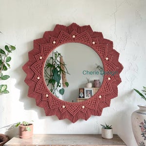 May include: A round wall mirror framed by intricate, dark terracotta-coloured macrame cord arranged in a sunburst pattern. The design features geometric knotting and small, light-coloured wooden beads embedded along the inner edge. The text "Cherie Design" is visible on the mirror surface.