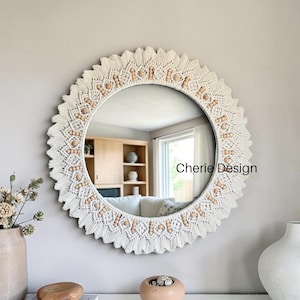 May include: A round wall mirror featuring an intricate off-white macrame border with a sunburst design. The frame is accented with natural light-colored wooden beads arranged in a circular pattern. The text Cherie Design is visible on the glass.