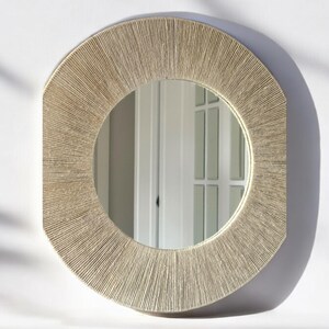 May include: A round wall mirror featuring a wide, textured frame wrapped in light beige natural fiber cord. The frame has a sunburst-like pattern created by the tightly wound vertical strands, surrounding a clear, circular reflective glass center.