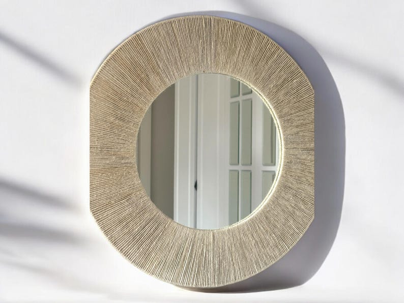 May include: A round wall mirror featuring a wide, textured frame wrapped in light beige natural fiber cord. The frame has a sunburst-like pattern created by the tightly wound vertical strands, surrounding a clear, circular reflective glass center.