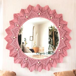 May include: A circular wall mirror framed by intricate dusty pink macrame cord arranged in a sunburst pattern with pointed edges and small round wooden beads integrated into the design. The text Cherie Design is visible on the mirror surface.