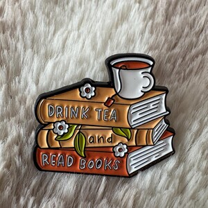 May include: A metal enamel pin featuring a stack of three books in shades of orange and brown. A white teacup sits on top. The text reads DRINK TEA and READ BOOKS. Small white flowers with brown centres and green leaves decorate the book spines.