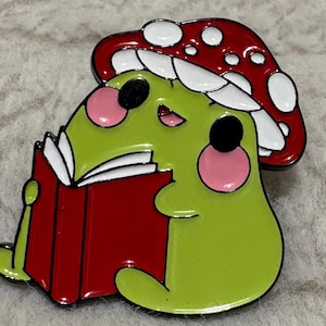 May include: A green frog-like character with pink cheeks and black eyes, wearing a red mushroom hat, is reading a red book. The enamel pin has a black outline and is set against a neutral background.