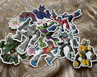 Könnte beinhalten: Ein Set mit neun Transformers-Charakteraufklebern mit verschiedenen Charakteren aus der beliebten Zeichentrickserie. Die Aufkleber sind auf weißem Papier gedruckt und in verschiedenen Farben gehalten, darunter Rot, Blau, Grün, Gelb und Schwarz. Die Aufkleber sind alle unterschiedlich groß und geformt und zeigen alle das Transformers-Logo. Der Text auf den Aufklebern lautet "Unsere Helden sind in Gefahr!" und "Das ist schlecht, Optimus!"